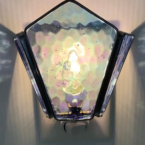 May include: A iridescent glass nightlight with a black metal frame in the shape of a pentagon. The nightlight is plugged into a standard electrical outlet.