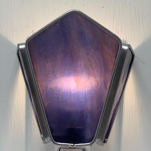 May include: A decorative night light with a geometric, pentagon-shaped shade. The shade features a blend of purple and pink hues, encased in a silver metal frame. The base is white and plugs into a standard electrical outlet.
