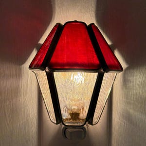 May include: A stained glass night light with a red top and clear, textured sides. The night light has a black frame and a warm, yellow light emanating from within. The design resembles a traditional lantern.