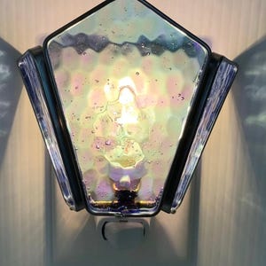 May include: A pentagonal plug-in night light featuring iridescent, textured glass panels with visible air bubbles. The glass is framed in dark metal, and the light emits a warm glow from within, casting soft shadows on the wall behind it.