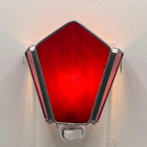May include: A pentagonal plug-in night light featuring vibrant red glass panels framed in silver-toned metal. The fixture includes a white plastic base with a small toggle switch at the bottom, designed to be inserted directly into a standard wall outlet.