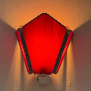 May include: A plug-in wall night light featuring a pentagonal shade made of vibrant red glass panels held together by dark metal soldering. The light emits a warm, glowing illumination from behind the red glass, casting a soft light.