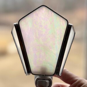 May include: A stained glass night light with a pentagon-shaped iridescent panel. The panel is framed with dark metal and flanked by two angled, rectangular panels. The night light has a standard plug.