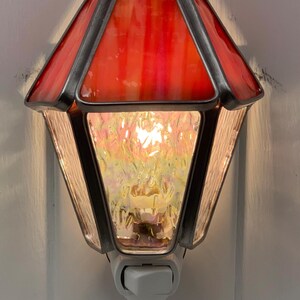 May include: A stained glass night light with a red and orange shade. The night light has a geometric shape with a textured, iridescent glass panel. The metal frame is dark, and the light is plugged into a white outlet.