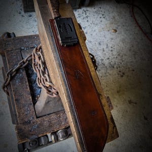 Op de afbeelding: Een handgemaakte bruin lederen schede met een mes. De schede heeft een donkerbruine afwerking en een gestempeld logo. Het mes heeft een getextureerde handgreep en een zwarte clip. De schede ligt op een houten oppervlak.