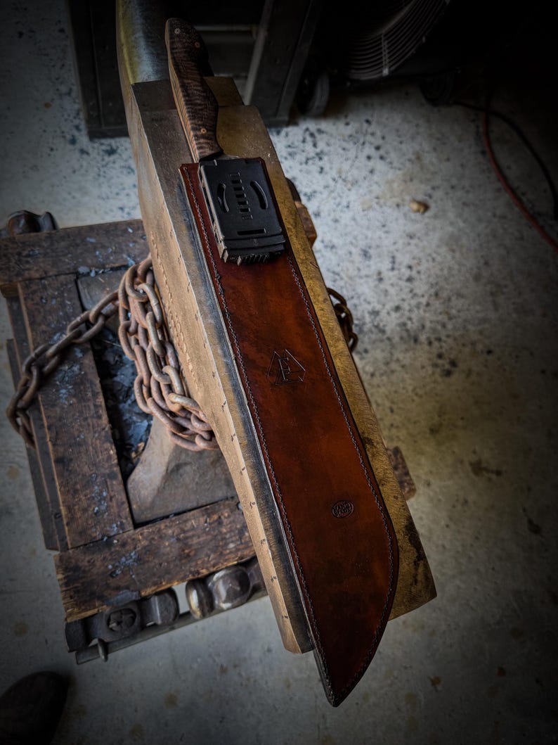 Op de afbeelding: Een handgemaakte bruin lederen schede met een mes. De schede heeft een donkerbruine afwerking en een gestempeld logo. Het mes heeft een getextureerde handgreep en een zwarte clip. De schede ligt op een houten oppervlak.