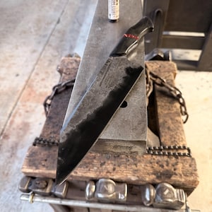 May include: A handcrafted, black chef's knife with a dark wooden handle and a red accent. The knife rests on a metal anvil, with a bottle of "FREEDOM LUBE" in the background. The blade has a dark, textured finish.
