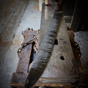 Op de afbeelding: Een groot, handgemaakt mes met een donker, getextureerd lemmet en een houten handvat rust op een metalen aambeeld. Het lemmet heeft een uniek patroon. Het aambeeld staat op een houten voet met een ketting.