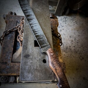 Op de afbeelding: Een groot, handgemaakt mes met een gebogen lemmet en een houten handvat. Het lemmet heeft een getextureerde, donkere afwerking en het handvat heeft een rijk houtnerfpatroon. Het mes rust op een metalen aambeeld, wat het vakmanschap laat zien.