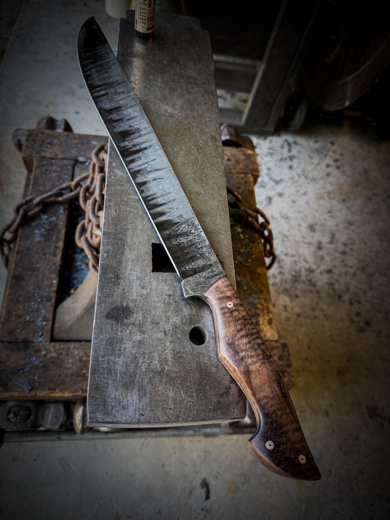 Op de afbeelding: Een groot, handgemaakt mes met een gebogen lemmet en een houten handvat. Het lemmet heeft een getextureerde, donkere afwerking en het handvat heeft een rijk houtnerfpatroon. Het mes rust op een metalen aambeeld, wat het vakmanschap laat zien.
