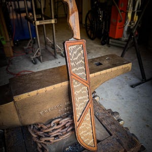 Op de afbeelding: Een handgemaakt mes met een houten handvat en een leren schede. De schede heeft een slangenpatroon en een bruine leren rand. Het mes ligt op een aambeeld, met een ketting eromheen.