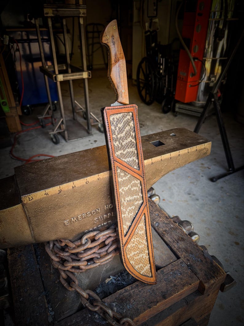Op de afbeelding: Een handgemaakt mes met een houten handvat en een leren schede. De schede heeft een slangenpatroon en een bruine leren rand. Het mes ligt op een aambeeld, met een ketting eromheen.