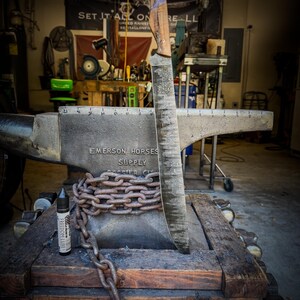 Op de afbeelding: Een groot, handgemaakt mes met een houten handvat en een getextureerd lemmet wordt getoond op een aambeeld. Een zware ketting wikkelt zich om de basis van het aambeeld. Een fles Freedom Lube staat op de houten basis. De achtergrond toont een werkplaats.