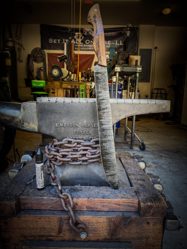 Op de afbeelding: Een groot, handgemaakt mes met een houten handvat en een getextureerd lemmet wordt getoond op een aambeeld. Een zware ketting wikkelt zich om de basis van het aambeeld. Een fles Freedom Lube staat op de houten basis. De achtergrond toont een werkplaats.