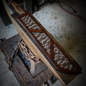 Op de afbeelding: Een handgemaakt mes met een houten handvat en een leren schede. De schede heeft een bruin en wit patroon, met bruine lederen accenten. Het mes en de schede worden op een houten oppervlak gepresenteerd.