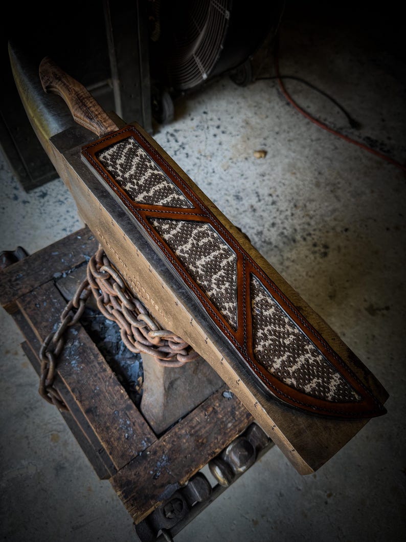 Op de afbeelding: Een handgemaakt mes met een houten handvat en een leren schede. De schede heeft een bruin en wit patroon, met bruine lederen accenten. Het mes en de schede worden op een houten oppervlak gepresenteerd.
