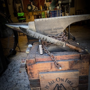 Op de afbeelding: Een groot, zilveren aambeeld met de tekst "EMERSON HORSESHOE SUPPLY" en een handgesmeed mes met een houten handvat rusten op een houten kist. Een flesje "Freedom Lube" ligt bij het mes. De kist heeft een houten bord met "SET IT ALL ON FIRE, LLC".