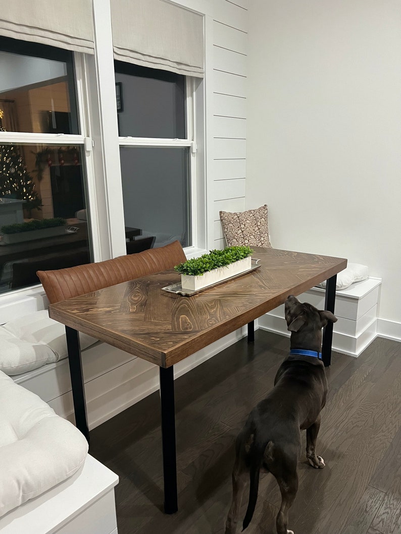 May include: A dark brown wooden table with black metal legs, a white cushion bench, and a black dog standing next to the table.