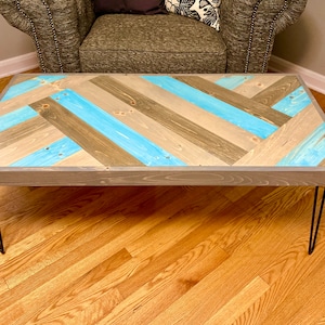 May include: A rectangular coffee table featuring a herringbone pattern on the tabletop, constructed from wooden planks in alternating shades of natural light brown, weathered gray, and bright turquoise blue, supported by four thin black metal hairpin legs.