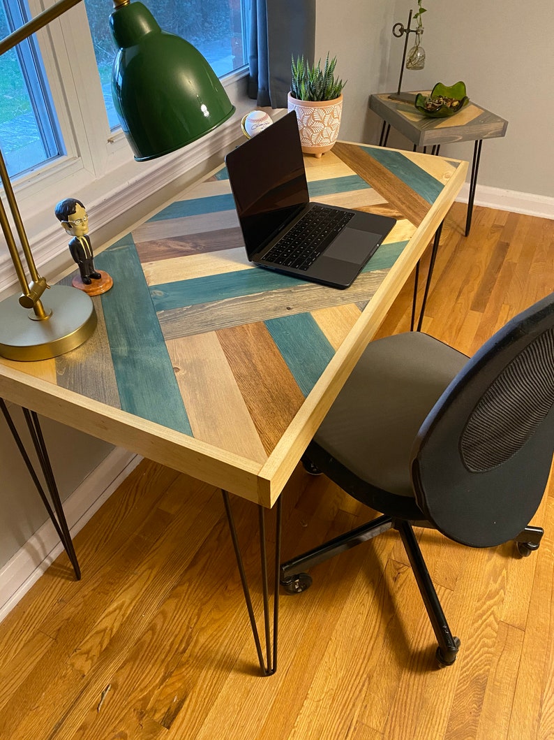 May include: A wooden desk with a chevron pattern top in shades of blue, brown, and beige. The desk has black hairpin legs and a laptop computer on the surface. A small side table with a gray top is visible in the background.