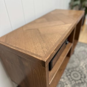 May include: A wooden media console with a light brown finish. The top features a chevron pattern, and the unit has open shelving. The console is designed for a living room or entertainment area.