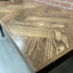 May include: A close-up of a wooden table top with a herringbone pattern. The table top is a dark brown color and has a smooth, polished finish.