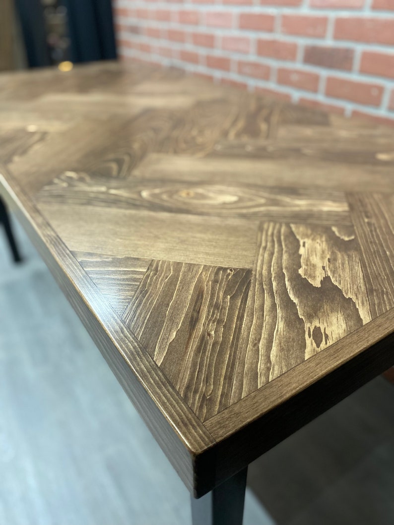 May include: A close-up of a wooden table top with a herringbone pattern. The table top is a dark brown color and has a smooth, polished finish.