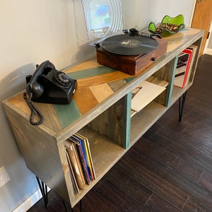 May include: A wooden record player stand with a chevron pattern top. The stand has three shelves and is painted in shades of gray, blue, and brown. It has black hairpin legs and a vintage rotary phone on the top shelf. A record player with a clear dust cover is on the top shelf, and a record is on the turntable. There are records on the bottom shelf.