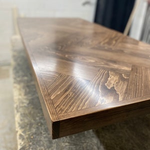 May include: Close-up of a dark brown wooden table top with a herringbone pattern. The table top is smooth and polished, with a slight sheen.