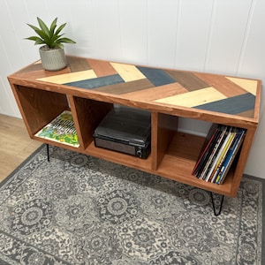 May include: A wooden media console with a multi-colored chevron pattern on top. The console has three compartments, one with a record player, one with vinyl records, and one with a magazine. The console is supported by black hairpin legs.