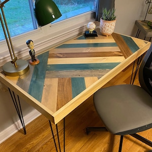 May include: A wooden desk with a chevron pattern top in shades of blue, brown, and beige. The desk has black hairpin legs and is sitting on a wood floor. A small figurine, a lamp, and a potted plant are on the desk.