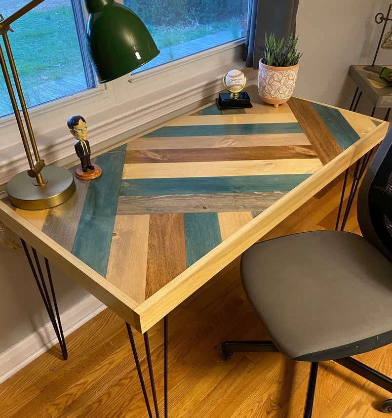 May include: A wooden desk with a chevron pattern top in shades of blue, brown, and beige. The desk has black hairpin legs and is sitting on a wood floor. A small figurine, a lamp, and a potted plant are on the desk.