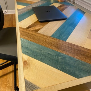 May include: A wooden desk with a chevron pattern in shades of blue, brown, and beige. The desk has a natural wood frame and a laptop computer is sitting on the surface.