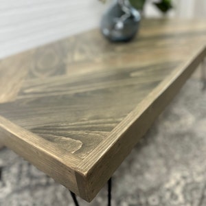May include: A wooden coffee table with a chevron pattern top and black hairpin legs. The table is stained a light gray color.