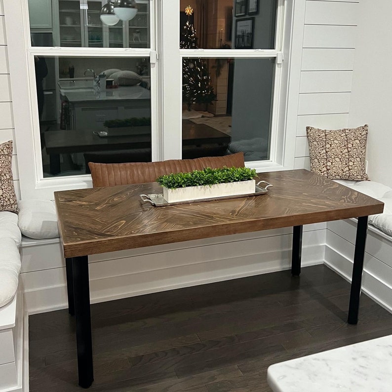 May include: A dark brown wooden table with black metal legs sits in a window nook. The table has a chevron pattern and is decorated with a small potted plant in a white planter. The table is surrounded by white cushions.