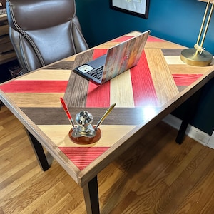 May include: A wooden desk with a chevron pattern in red, brown, and beige. A laptop computer with a patterned case is on the desk. Two pens are in a silver pen holder.