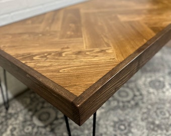May include: A wooden table with a herringbone pattern top and a dark brown border. The table has black hairpin legs.