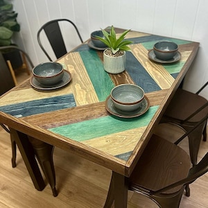 May include: A rectangular dining table with a multi-colored, chevron-patterned top. The table's surface features alternating wooden planks in shades of brown, green, and blue. Four matching bowls and saucers are set on the table, along with a small potted plant. The table is supported by dark metal legs.