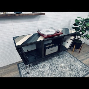 May include: A black and wood-toned media console with a geometric design on top. It features a red turntable, a Marshall speaker, and space for vinyl records. The console sits on black hairpin legs, and the room has a patterned rug.