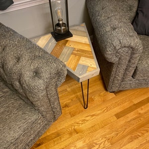 May include: A gray chevron patterned wooden side table with a black metal lamp and a gray armchair. The table has hairpin legs and the lamp has a glass shade.