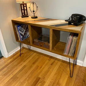 May include: A wooden media console with three open storage compartments, supported by four black metal hairpin legs. The top surface features a geometric inlay pattern of wood and dark resin. A black rotary telephone sits on the right side.
