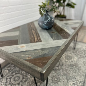 May include: A wooden coffee table with a chevron pattern top in shades of gray, brown, and white. The table has black hairpin legs.