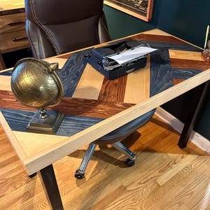 May include: A wooden desk with a chevron pattern in black, brown, and natural wood. The desk has a black metal frame and is topped with a vintage typewriter, a globe, and a pencil holder.