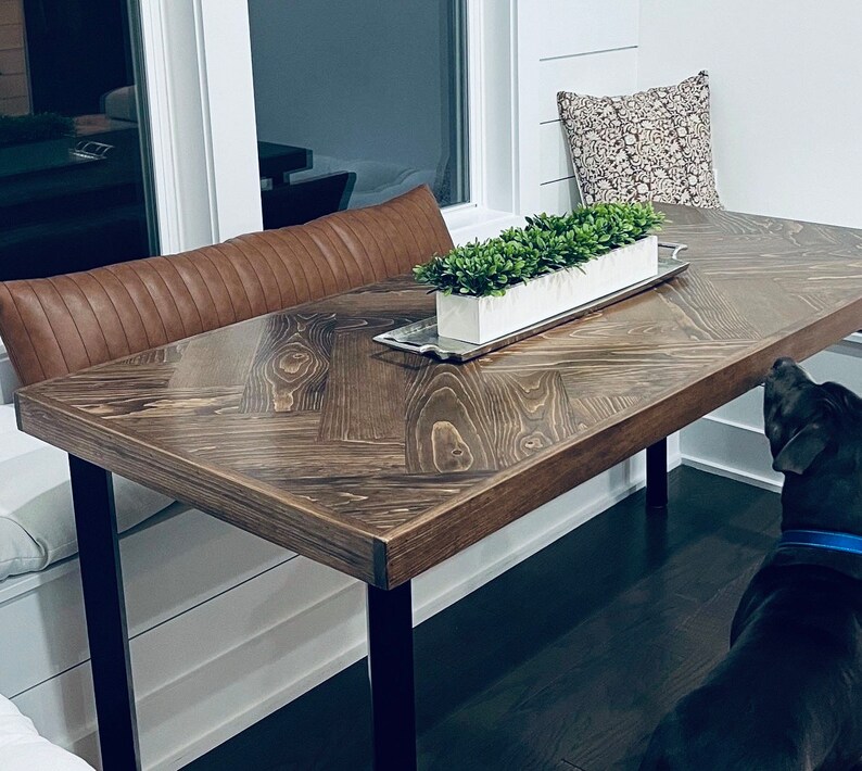 May include: A dark brown wooden table with a chevron pattern top and black metal legs. The table is set against a white wall and has a brown leather cushion bench on one side. A small potted plant sits on the table, and a black dog is in the foreground.
