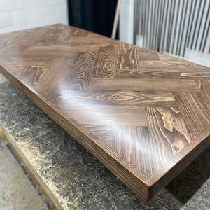 May include: A close-up of a dark brown wooden table top with a herringbone pattern. The table top is smooth and polished, with a slight sheen.