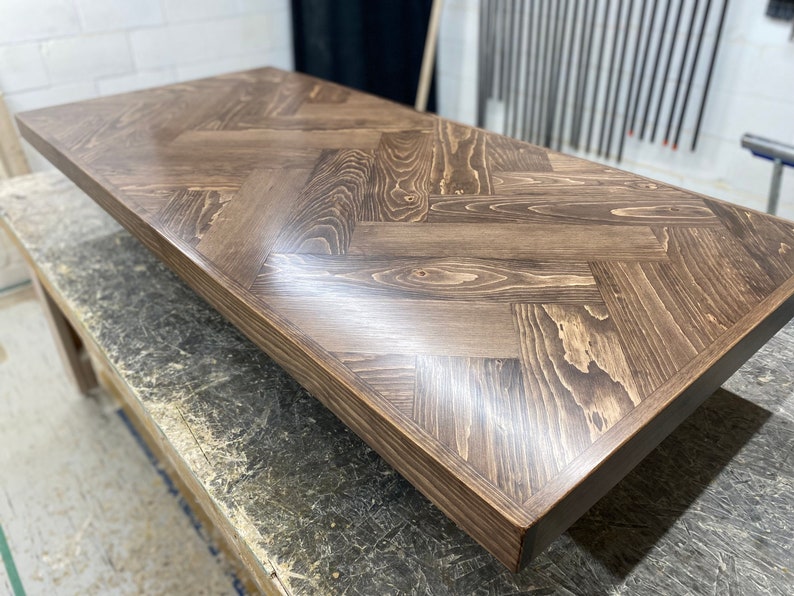 May include: A close-up of a dark brown wooden table top with a herringbone pattern. The table top is smooth and polished, with a slight sheen.