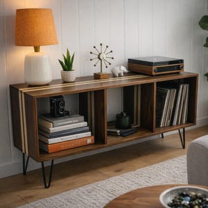 May include: A mid-century modern style media console made of dark wood with light wood accents. The console has three open compartments, black hairpin legs, and is topped with a record player, lamp, and decorative items. The console is filled with books and vinyl records.