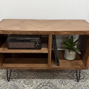 May include: A wooden media console with a herringbone pattern top. The console has two shelves and a compartment for a record player. A small potted plant and a book are visible. The console sits on black hairpin legs.