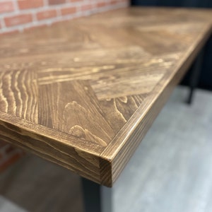 May include: Close-up of a dark brown wooden table top with a chevron pattern. The table top has a slightly rounded edge and is made of reclaimed wood.