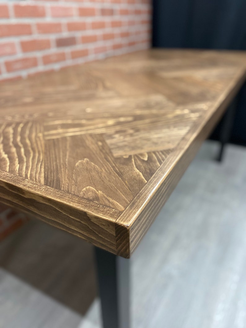 May include: Close-up of a dark brown wooden table top with a chevron pattern. The table top has a slightly rounded edge and is made of reclaimed wood.
